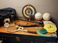 Overview of lot on tabletop showing bow, racquet, discs, dartboard, soccer balls, racquetball cans, duffel bag