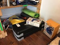 Group of fitness items under shelf: step platform, yoga mat, foam roller, resistance band, stability ball box, yellow boxed item