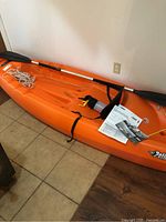 Full view of orange Pelican kayak with paddle, rope and manual on floor