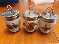 Three egg coddlers standing with lids on showing motifs