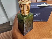Full view of green textured Lampe Berger lamp with gold crown cap and box behind