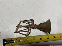 Front view of brass hand bell with scales handle beside tape measure showing length