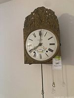 Front view of wall clock showing ornate brass surround, enamel dial, pendulum rod