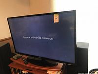 Front view of Samsung TV powered on displaying welcome screen with remote on stand