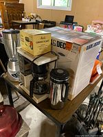 Table with boxed Black & Decker breadmaker, blender, coffee maker, thermos and boxed carafe