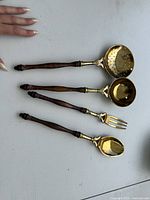Four large brass and wood decorative serving utensils laid out: slotted ladle, ladle, fork, spoon
