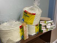 Overall lot showing stacked unopened packs of plates, boxed plate packs, and birch cutlery box on shelf
