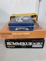 Stack showing Rummikub, Fox & Geese, yellow domino box, blue domino tin