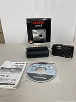 Both cameras boxed/present, accessories visible (case, Lumix camera, Pentax box, manual, software).