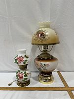 Both hurricane lamps side by side showing size difference, floral glass globes, brass-tone bases