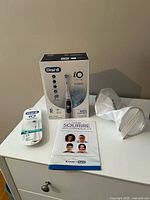 Front view of sealed Oral-B iO Series 6 toothbrush box, replacement brush head pack, booklet, pouch and plastic accessory on dresser