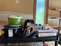 Shelf with all eight items: Crock-Pot, salad spinner on top, Frigidaire air fryer, Homasy diffuser box, Starfrit mandoline box, Salton food sealer, burger press, corkscrew partly visible