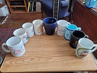 Group shot of all nine assorted mugs on tabletop, showing front designs and colour variations