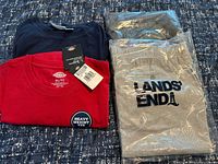 Four folded clothing items: red Dickies tee, navy tee, two bagged Lands' End grey shorts
