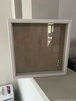 Front view of white shadow box showing glass front, beige backing and three push pins