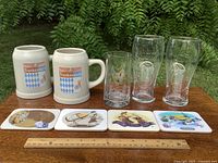 Front view of entire lot: 2 stoneware steins, 1 tumbler, 2 pilsner glasses, 4 coasters, ruler for scale