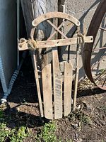 Front view of full sled leaning against post showing wood slats, faded lettering, metal runners, rope lashings