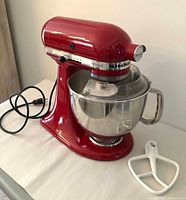 Left-front view of red KitchenAid Artisan mixer with stainless bowl and flat beater nearby