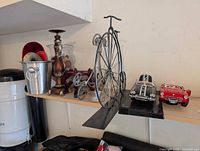 Shelf view showing ice bucket, candlestick, glass jar, wooden stand, miniature tricycle, penny-farthing sculpture and two model cars