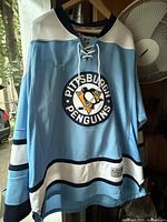 Front view of light blue Penguins jersey hanging, showing crest, stripes, lace-up collar