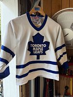 Front view of white Toronto Maple Leafs jersey hanging, shows logo and stripes