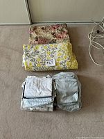 All bedding items folded and stacked: floral quilt on top of patterned duvet cover, two folded sheet sets and light grey pieces