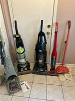 Lineup of five appliances against wall: two Bissell units, Dirt Devil upright and stick, Hoover polisher