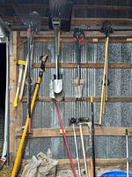 Assorted long-handled tools hanging: shovels, rakes, pitchfork, Fiskars telescopic pruner, splitting maul, pickaxe visible