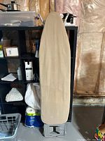 Full view of ironing board with beige cover leaning against shelving