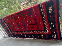 Full length view of kilim rug hanging over railing showing red field, geometric motifs, navy/black border and fringe