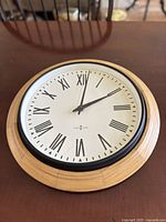 Front view of round wall clock showing wood frame, white face with Roman numerals