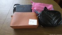 Four crossbody handbags arranged on tabletop: brown Nanette Lepore, black Steve Madden, black Herald, pink International Concepts with chain strap.