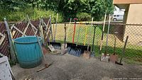 Overall lot with barrel and assorted shovels/rake lined along fence