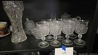 Group view showing vase, six goblets and other glass pieces on shelf