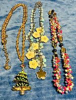 All three necklaces laid out: gold-tone chain with Christmas tree pendant, silver-tone floral charm necklace, braided beaded necklace