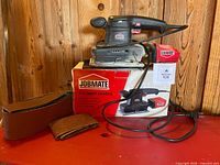 Sander with dust bag on top of box, power cord visible, sandpaper sheets nearby