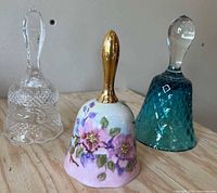 Group shot of three bells: clear pressed glass, teal glass, porcelain with gold handle