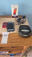 Group shot of all electronics on table: LG smartphone, red flip phone, Panasonic CD player, LG Aristo 4+ box, blue earbuds