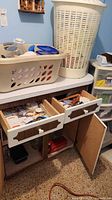 Counter with beige laundry basket containing containers, towel; open drawers with household items; tall white hamper beside