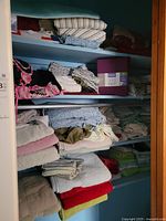 Closet shelves filled with folded linens: sheets, towels, blanket, crochet throw, packaged sheet set