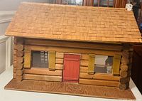 Front view of wooden log-cabin dollhouse showing cedar shingle roof, red door and window openings