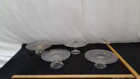 Four clear pressed glass pedestal cake stands on black surface with rulers for scale