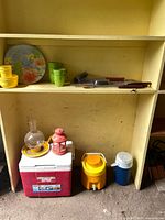 Shelved view showing plate, plastic drinkware, bbq tools, coolers, lanterns, lamp