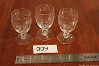 Three identical etched crystal stemmed glasses on wood surface with measurement scale and lot card