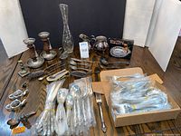 Overall lot view on table showing flatware bundles, serveware, candlesticks, vase, boxed tray
