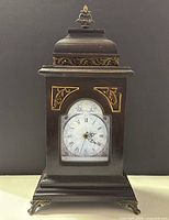 Front view showing clock face, wood case, gold accents, and feet