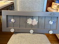 Full view of gray wood wall rack showing three ceramic hooks and floral scripted panel