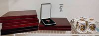 Stack of six red wooden display boxes, open presentation case with medal, two lidded mugs on right