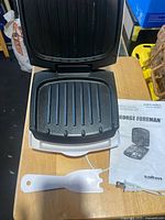 Open George Foreman grill showing cooking plates with manual and spatula beside