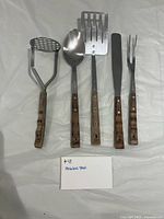 Front view of all five stainless steel utensils with wooden handles laid flat on surface